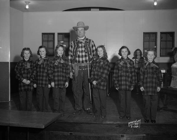 Group portrait of three sets of girl twins (Jean and Janette Heinz, Julia and Elizabeth Kalscheur and Arline and Irene Ripp), wearing plaid shirts, and Mr. Ripp wearing a cowboy costume. A woman musician is seated at a music stand in the background. Photograph taken at a Dane County Farm Bureau program at the Evangelical Lutheran Church.