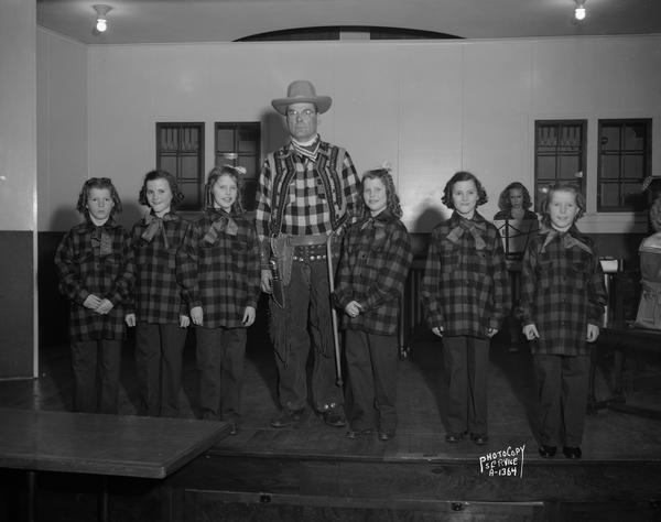 Group portrait of three sets of girl twins (Jean and Janette Heinz, Julia and Elizabeth Kalscheur and Arline and Irene Ripp), wearing plaid shirts, and Mr. Ripp wearing a cowboy costume.  A woman musician is seated at a music stand in the background.  Photograph taken at a Dane County Farm Bureau program at the Evangelical Lutheran Church.