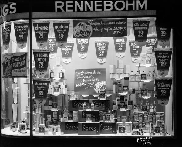 Display window of an unidentified Rennebohm Drug Store featuring a wide variety of common drug store merchandise.  Sign says "Shoppers who are smart and wise always buy the larger size."  Display shows the prices for all the merchandise displayed.
