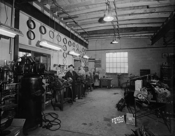 Three workers standing beside their machines in the machine shop at Winnebago Auto Replacement Company, 103 North Park Street.