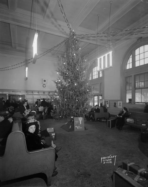 Large Christmas tree in the waiting room of the Milwaukee Road Railroad Station, 644 West Washington Avenue, with passengers and the sign: "Give War Bonds, the present with a future."
