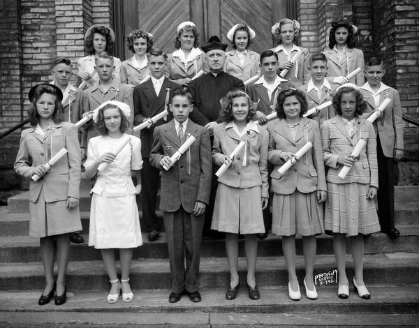 Group portrait of boy and girl confirmation group, and a priest, standing on the steps at St. Patrick's Catholic Church, 410 East Main Street.