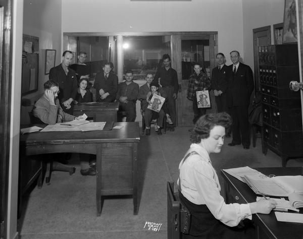 Office interior of the Madison News Agency, 446 West Gilman Street, taken from entrance. Employees are sitting in the room, and in the background a group of men and children are standing and sitting.