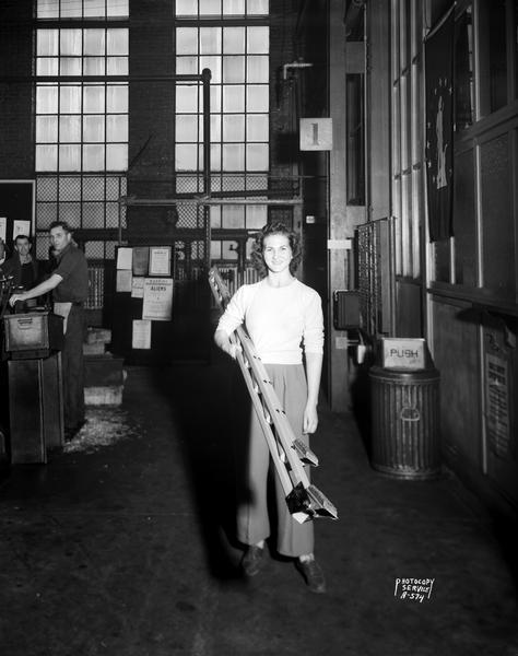 A woman carrying a folding ladder on the Madison-Kipp Corporation factory floor.
