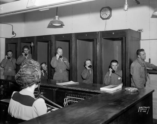 Men, some of whom are dressed in overalls, make phone calls in a bank of telephone booths at Truax Army Air Field in the recreation hall and switchboard area. A woman wearing a telephone headset is sitting in the foreground.