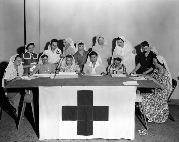 State female employees making surgical dressings / bandages for the American Red Cross in the Wisconsin State Employees Association building, 448 W. Washington Avenue. Seated, l to r: Marion Block, Lenore Hassett, Caroline Dagestad, Lydia Stumpf (leader), Vyrgil Tuft, and Dorothy Ryan. Standing, l to r : Lenore Brownlee, Rosaline Bee, Eulalia Fader, Eleanor DeMerse, Rosella Marshall, Zenobia Peachey and Dorothy Wilhelm.