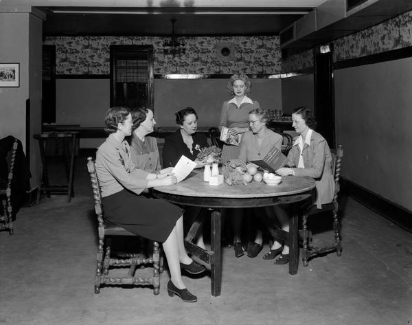 Group of women reading "Canteen Corps" pamphlets and planning Red Cross nutrition classes for civil service employees. l to r: Vivian Holland, chairman of nutrition and canteen enrollment in the Capitol and State Office Building; Ruby Taff, Mendota State Hospital chairman; Alice Ferguson, teacher of Civil Service nutrition classes; Janet Nelson, (standing) Wisconsin State Employee Magazine reporter; Mrs. O.A. Fried, chairman of Red Cross nutrition and canteen work in Dane County; and Carol Miller, University Employees nutrition chairman. Classes were formed in response to young men being rejected from military service on the grounds of health deficiency.