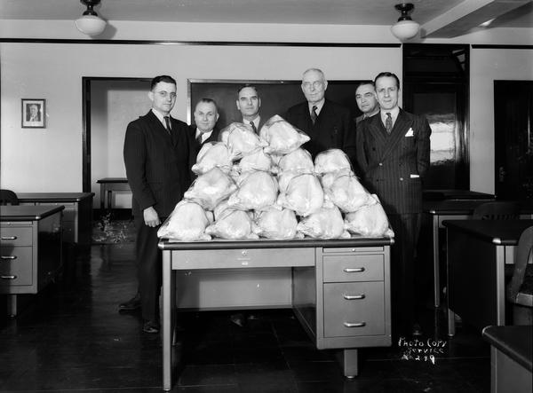 Six men stand behind a desk piled with fifteen dressed and wrapped turkeys.