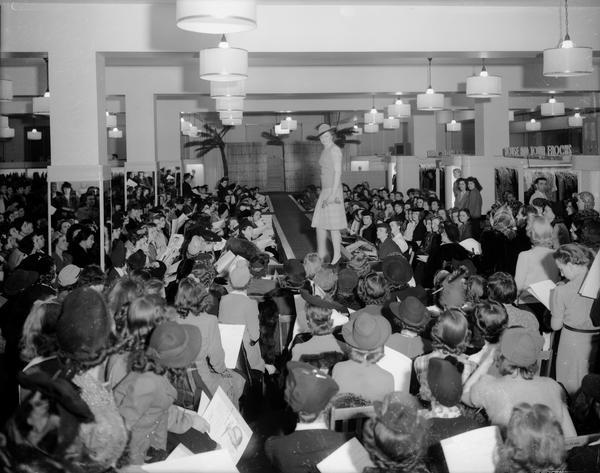 Women's style show with female model on runway with women seated in chairs on main floor at Manchesters Department Store.
