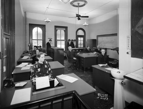 Interior view of Dane County Title Company office, 109 S. Fairchild Street, with 11 people, some of them using typewriters. A water fountain (bubbler) is along the wall on the right.