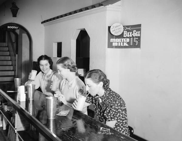 Three women are drinking malts at the counter of McCoy's Ice Cream Parlor, 507 State Street. A sign on the wall reads: "A Balanced meal in every glassful! Drink Bee-Gee's Malted Milk, 15 cents."