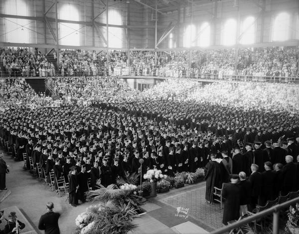 Panoramic view of University of Wisconsin graduation exercises, taken from behind the podium in the Field House. Graduates are standing on the floor, and the audience is observing from the bleachers.