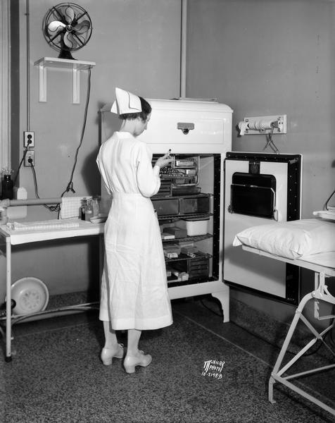 A nurse in uniform standing in front of an open refrigerator, checking vials of medicines at the Wisconsin General Hospital.