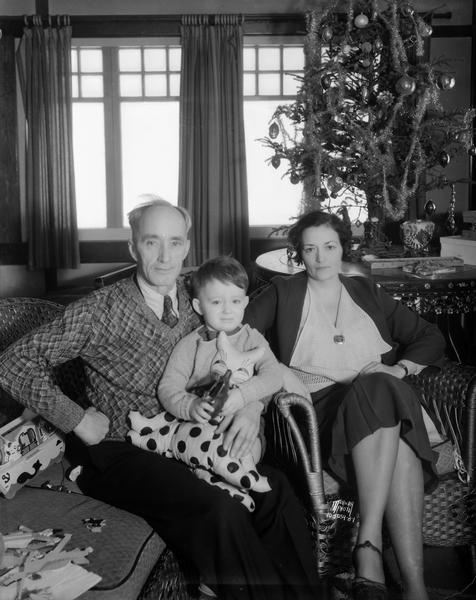 Group portrait of William J. and Mae Hobbins, and their son Jimmie sitting on a sofa in front of a Christmas tree in their home near Camp Indianola. Hobbins was the president of the bankrupt Capital City Bank. He was convicted of fraud and pardoned by Governor Schmedeman on Christmas Day.