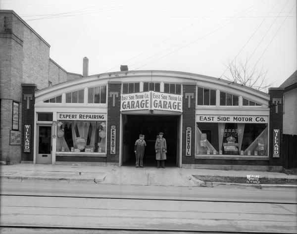 Two men are standing in the doorway of the East Side Motor Co. garage, 2305 Atwood Avenue, featuring Willard storage batteries, Pennzoil, and Eveready and Prestone.