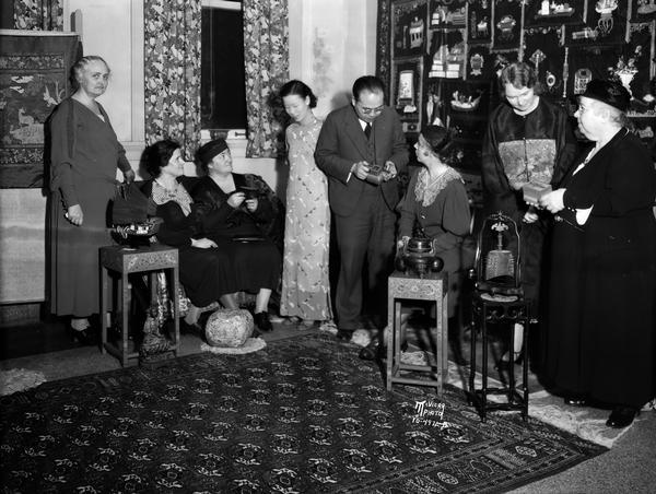 Group of men and women viewing Chinese art and artifacts including tables, bell, teapot and Chinese junk with sails. On the wall in the back are pictures of other Chinese artifacts. Several philanthropies of the Madison Woman's club will benefit from the proceeds of the afternoon bridge and art exhibit held at the club.