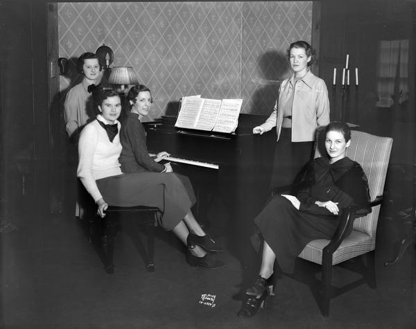 Group portrait of five "best dressed co-eds" gathered around a piano, at the University of Wisconsin. They were picked in a poll conducted by the "Daily Cardinal." All are sorority women.