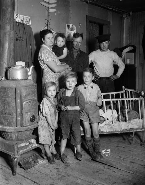 Portrait of seven of the twelve members of the Chris Copus family standing beside a pot belly stove in their makeshift garage home on Roth Street. They were denied relief by Dane County during the Depression.