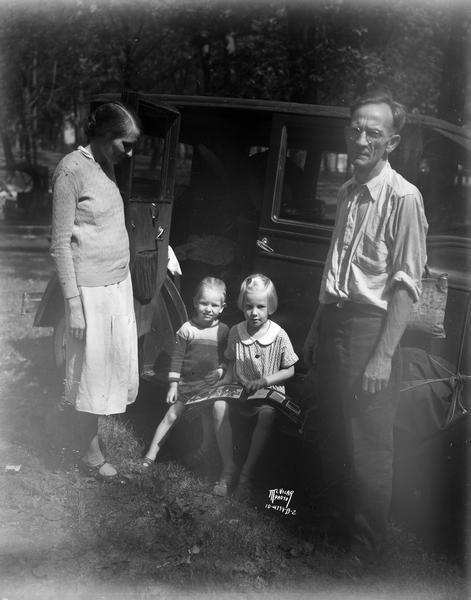 Edward O'Connell (General Pershing's chauffeur), and wife standing beside an automobile, with two children sitting on the automobile's running board and looking at a book.
