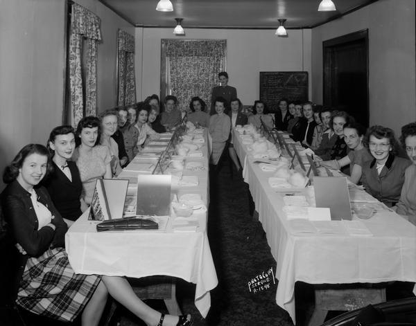 Twenty-four women seated in front of individual mirrors on two tables ready for instruction about Cara Nome cosmetics. Sign says: "Knowledge, Work, Personality equals profit."