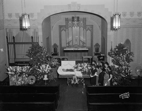 Elevated view of Walter Becker's body in a casket in the front of the Zion Evangelical Lutheran Church, 247 Division Street. The casket is surrounded by floral bouquets, and his relatives are standing on the right looking on. He was born in Germany, worked for Fuller & Johnson, Steinle & Gisholt and was the superintendant of buildings and grounds for the church for 20 years.