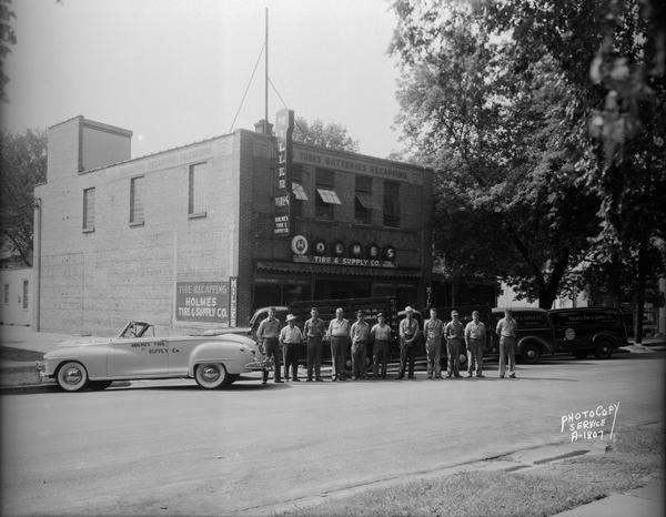 Line-up of four trucks and one car in front of Holmes Tire and Supply Company, 431 West Main Street, with eleven employees.