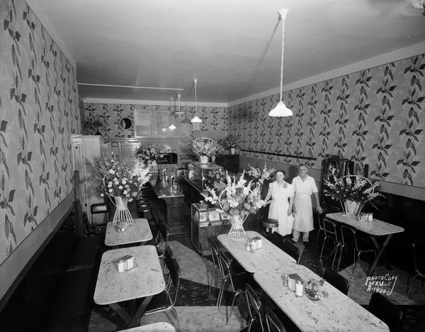 Slightly elevated view of the interior of Young's Cafe, located at 316 East Wilson Street. Joseph and Anna Young are standing in the Cafe surrounded by bouquets on opening day.