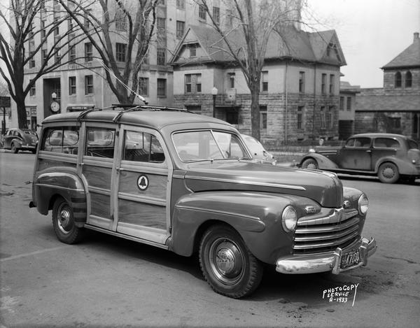 Ford "Woody" station wagon test car with antenna for mobile telephone, on West Washington Avenue in front of the Grace Episcopal Church. Also included in the view is Cornelia Vilas Guild Hall, The Old Rectory Gift Shop, 16 West Washington Avenue, and Wisconsin Power and Light Company building.