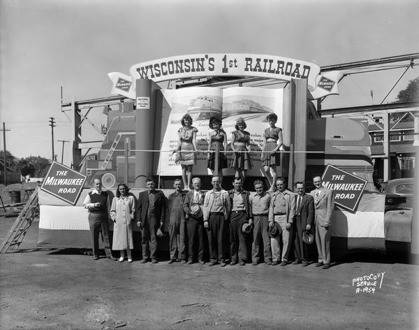 Milwaukee Road float for the Wisconsin Centennial Parade, "Wisconsin's First Railroad," with women on the float and eleven workers standing in front of the float.