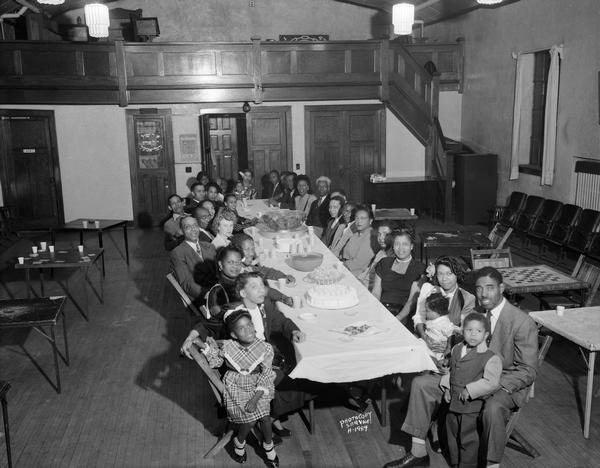 Slightly elevated view of a group of African American families sitting along a long table, celebrating a birthday party at the Workmens Circle Clubhouse, 43 North Mills Street.