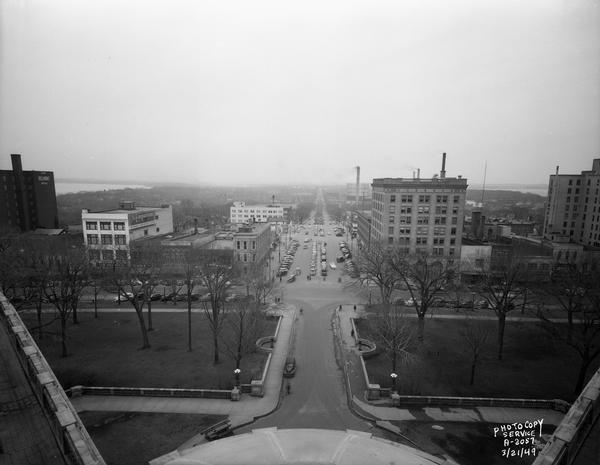 Elevated view from the Wisconsin State Capitol, showing Pinckney Street side of the Square, looking down East Washington Avenue. Buildings include the Belmont Hotel, Emporium, American Exchange Bank, Ray-O-Vac Office, First National Bank and Tenney Building. Lake Mendota is in the far background on the left, and Lake Monona is on the right.