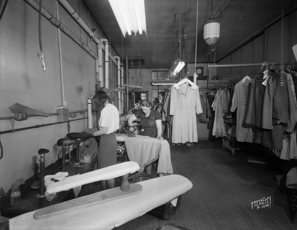 Interior of Four Lakes Cleaners, 612 South Park Street, with work room showing two women pressing clothes, and racks of clean clothes.