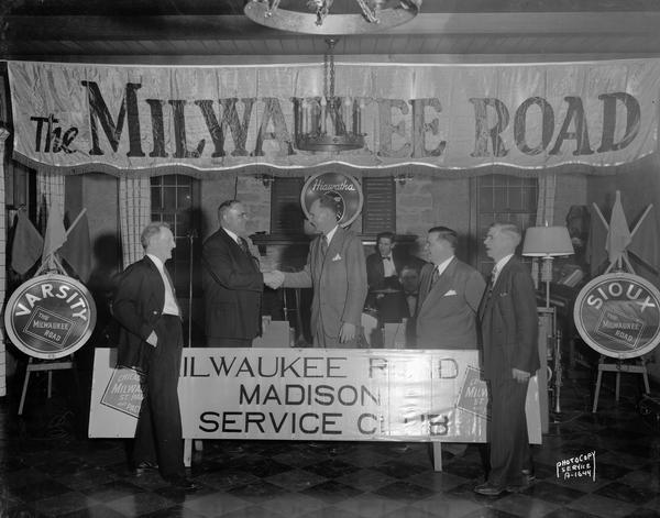 Milwaukee Road Service Club officers greeting superintendent at party given at Nakoma Country Club, with Milwaukee Road Service Club sign and banner and Varsity and Sioux train signs.