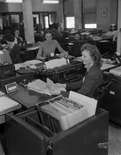 Close-up of a woman sitting in front of a Remington Accounting Machine. In the background are other women and a man in the office using other office machines, at CUNA (Credit Union National Association).