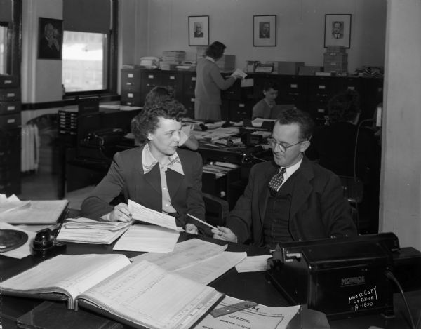 Man and woman reviewing a document in an office, with three women working in the background at CUNA (Credit Union National Association).