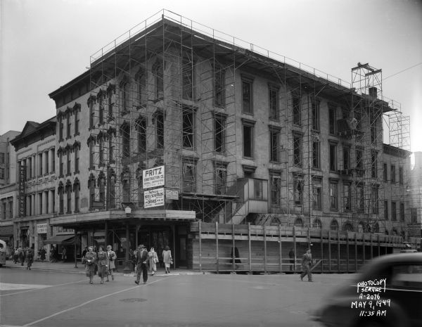 View from Park Hotel towards the Blied Building, 29 West Main Street and Carroll Street facade, under construction. The corner of the building is covered with construction scaffolding. Western Union is at 21 West Main Street, Madison Bank and Trust at 23 West Main Street, Walk Over Shoe Store at 25 West Main Street, Forbes-Meagher Music Store at 27 West Main Street. There are pedestrians crossing the street.