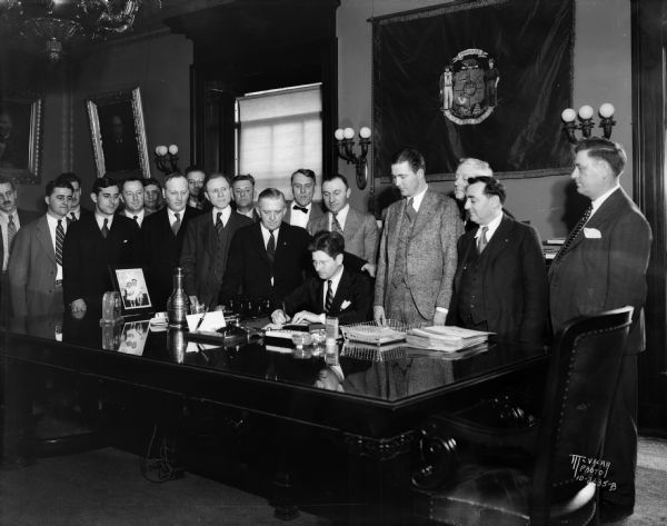 Governor Philip La Follette signing old age pension bill surrounded by interested parties. Assemblyman Oscar Schmiele stands at the governor's left shoulder, Senator Walter Polakowski is at the extreme right, Everett C. Gerry, Sec. Eagles Club at extreme left. Senator Leonard Fons is third from the left. Other men are Eagles who lobbied for the old age pension bill.