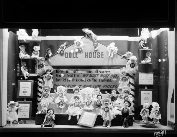 Hill's Department Store, 202 State Street, window display of dolls and dollhouse in the background. "We've been sitting around all summer, Now the heat is on, We Must Move Out, Don't let us sit around on the shelves, Buy us now at new low prices."