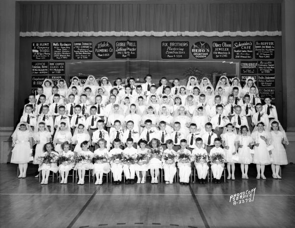 St. Bernard's Roman Catholic Church, 2460 Atwood Avenue. Group portrait of a large First Communion class in front of the stage. Includes business advertising signs on a screen in the background.
