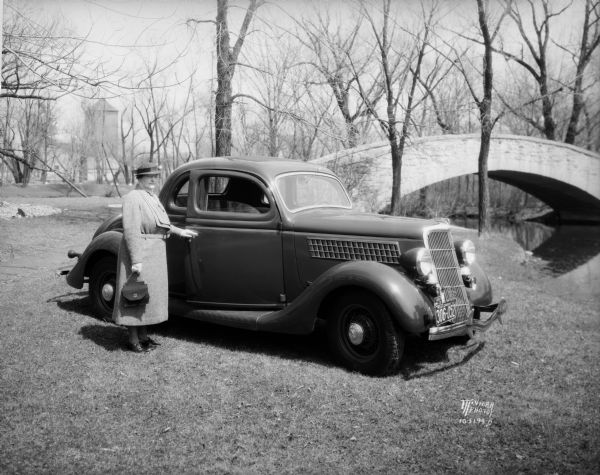 Miss Pyre standing next to a Ford V-8 coupe near a bridge at Tenney Park. A building with a tower is in the background, perhaps the Hausmann Brewing Company Malt House.