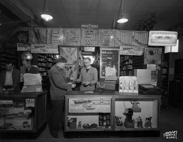 Interior of Richard Ela Company, 744 Williamson Street, showing a sales counter with a large display of tools. Three clerks and one customer are standing near the counter.