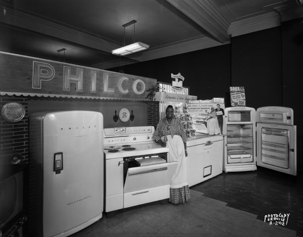 Woman dressed as Aunt Jemima standing with merchandise display, at the Casey and O'Brien Store, 124 West Mifflin Street, featuring Philco appliances including two refrigerators, a chest freezer, and a stove.
