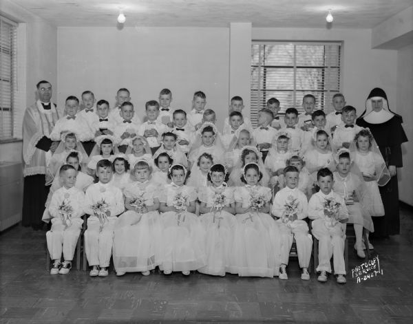 A group portrait of children in a First Communion class, with a priest and nun, of St. John the Baptist Catholic Church.