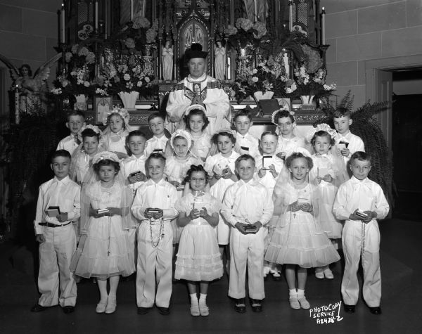 A group portrait of a First Communion class taken in front of the altar, along with a priest, inside the St. Peter Catholic Church.