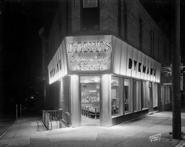 Exterior night view from street of Dykman's restaurant, located at 107 West Main Street. The door is open and stools are along a counter.