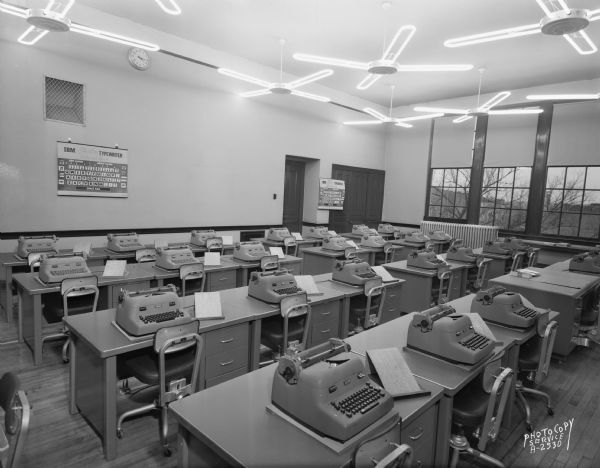Madison Vocational and Adult Education School, 211-213 North Carroll Street. View of typing room #442 from the back, featuring IBM typewriters. The ceiling fans appear to have neon lighting along the blades. For Haskell Desk Co.