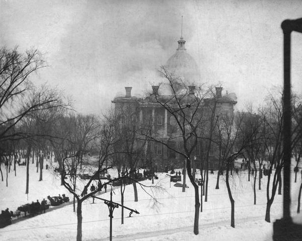 Fire at the third Wisconsin State Capitol, showing the South Wing, and spectators gathered on the path. Some of the salvaged contents of the building are scattered on the snow.