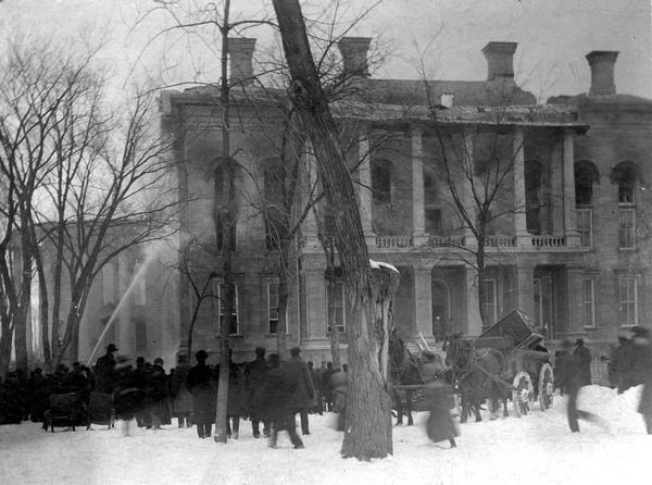 Wisconsin State Capitol fire, showing a crowd watching near the South Wing. Some of the contents of the building are already in wagons ready to be hauled away.