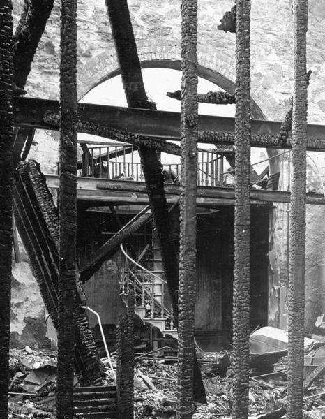 Ruins of the second floor of the North Wing (looking south), after the fire that destroyed most of the third Wisconsin State Capitol. In addition to the charred timbers, this close-up reveals some of the decorative details of a metal circular staircase. The specifications for the 1882 addition provides additional information: "All stair rails to be made of oak moulded and grooved over the iron band, and secured firmly thereto; to be one piece for each flight; the oak to be thoroughly kiln dried and must be free from knots, checks or cross grains."