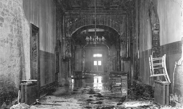 A close-up of the damage caused to the third Wisconsin State Capitol by the fire of February 26-27, showing the first floor, and looking west through the Rotunda toward the exit. Close scrutiny of this view reveals some details of the original appearance of the Rotunda (compare this to Image ID: 7074), but most notable in the Rotunda is the presence of "Genius of Wisconsin," a female figure with an eagle on her shoulder. This marble statue by Helen Farnsworth Mears is one of the few objects from the third Wisconsin State Capitol on display in the present Capitol. You will find "Genius" one floor above ground level at the southeast entrance.